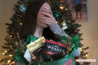 A teenage girl in front of Christmas tree holding a gift bag with 4 bags of jerky.  Holding her hand over her mouth in excitement. 