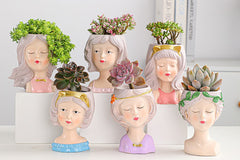 A collection of six small, ceramic girl-head succulent planters arranged on white display blocks, each containing a different plant