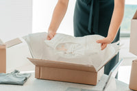 Woman wearing a black dress is pictured. She is grabbing a white sweater from a cardboard box. Box is sitting on top of a table. 