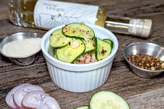 A spicy cucumber salad in a white bowl, surrounded by its ingredients: ramp vinegar, sugar, crushed red pepper, shallot, and cucumber.