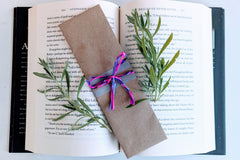 A fantasy romance-themed bookmark wrapped in brown craft paper and adorned with pink and purple ribbons rests on an open book.