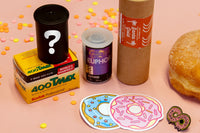 4 rolls of film as well as two stickers of a donut and a pin that says Stay Hungry sit on a pink table