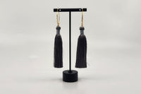 Elegant tassel earrings by LOKA.HAUS, with intricate gray tassels suspended from gold chains, displayed on a sleek black stand.