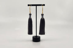 Elegant tassel earrings by LOKA.HAUS, with intricate gray tassels suspended from gold chains, displayed on a sleek black stand.