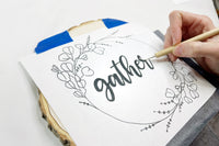 Close up view of transferring a woodburning pattern that says "Gather" to a slice of wood using a tracing tool and carbon paper.