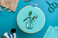 Embroidery project featuring green plant cuttings in water surrounded by various embroidery kit supplies.