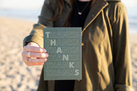 a female at the beach holding out a greeting card that says THANKS on it with the word thanks repeated over and over