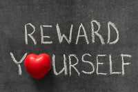 A chalk board with the words REWARD YOURSELF written in capital letters with white font. The letter O is replaced with a red heart.
