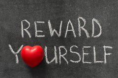 A chalk board with the words REWARD YOURSELF written in capital letters with white font. The letter O is replaced with a red heart.