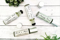 Four essential oil rollers on white washed wood with plants. Lights Out, Rise + Shine, Happy Place, and Bust a Move.