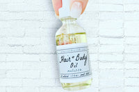 Two fingers holding the Melissa Hair + Body Oil by Greenmade goods in front of a white brick wall showcasing dried flowers inside the bottle