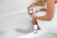 woman sitting on the edge of the bath tub with legs in the full tub holding a whipped soap scrub and wearing a white bath towel