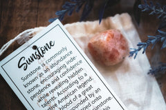 orange stone on a cream muslin bag with a white hang tag with black writing all next to a purple flower on top of a wood table