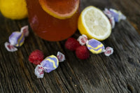 Raspberry Lemonade Saltwater Taffy is nestled between fresh raspberries and lemons with a large glass of ice-cold lemonade. 
