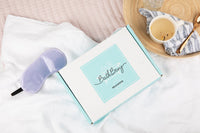 closed Bath Bevy Subscription box next to purple silky sleep mask and cup of tea with pink and white textile background