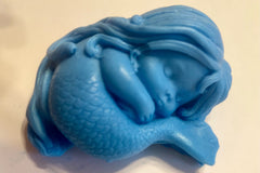 A shiny blue soap shaped like a mermaid sleeping on her own tail reflects light and rests on a white base.