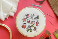 Embroidery project surrounded by kit supplies. Project features kettles, cups, and plant cuttings stitched onto off white fabric.