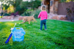 Remedy application bag and attached sprayer shown in grass with child and pet behind it. Remedy spray is pet- and child-friendly.