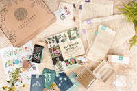 A closed Earthlove subscription box surrounded by a plant book, packets of seeds, and other gardening supplies.