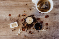 The image looks down on an open jar candle. Coffee beans and unlit matches are scattered around the base of the candle jar. 