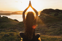 a rocky scenary facing a body of water. Sun is low in the sky. A female with long hair seems to be meditating while sitting.