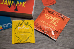 Corner of book and two tea bags, all orange and yellow themed. Blue Queerly Curated bookmark peeks out on left side