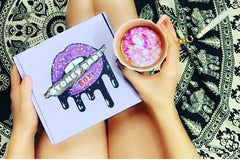 A woman holding a Stoney Babe subscription box on her lap and a mug of some sparkly, pink and white substance.