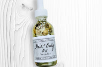 A bottle of the Lavender Hair + Body Oil by Greenmade goods on white wood showing dried lavender and jasmine buds inside the bottle.