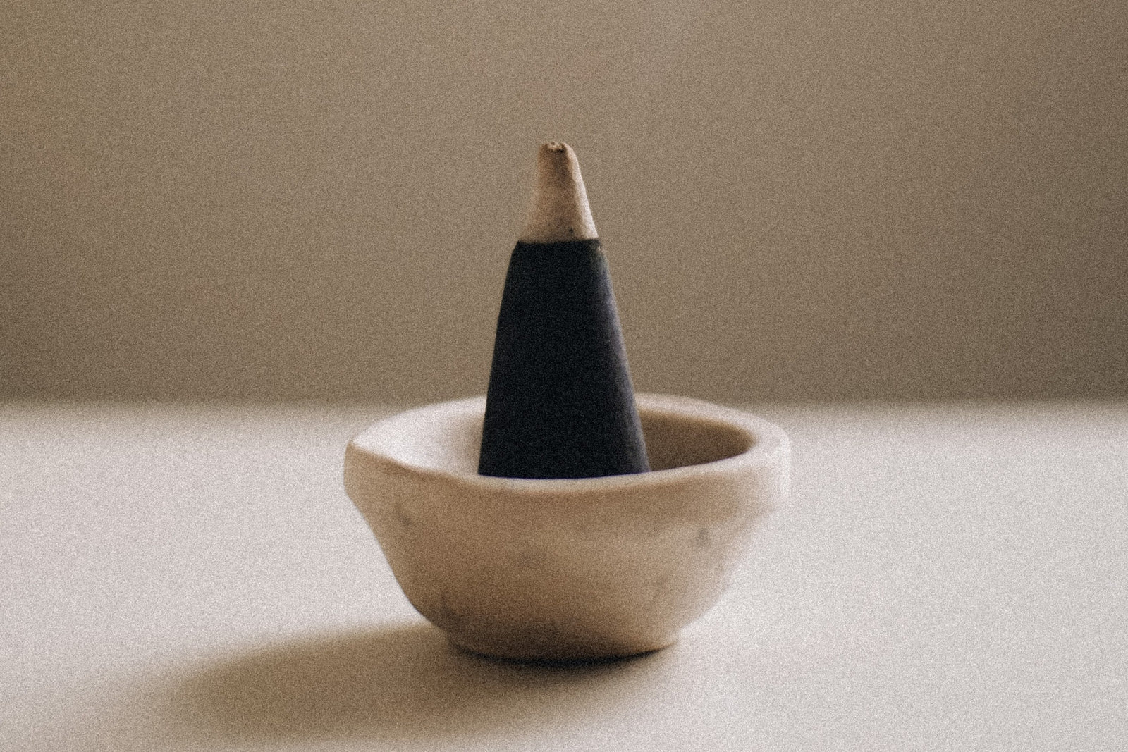 white table with small ceramic beige bowl with black cone incense with grey tip there is a darker beige wall