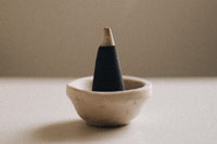 white table with small ceramic beige bowl with black cone incense with grey tip there is a darker beige wall