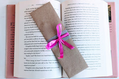 A Romance-themed bookmark is wrapped in brown craft paper and decorated with pink ribbon. It rests on an open hardback book.