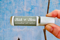 Two fingers holding the Stick + Stone Essential Oil roller bottle with an white washed blue concrete wall in the background.
