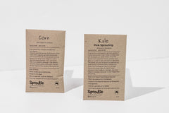 Two seed packets sit upright on a white background. They are brown paper. One says "corn" and the other "kale, pink sprouting".