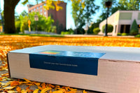 Queerly Curated box (white box, blue label with logo) laying on the ground in orange leaves, in a small-town city scape.