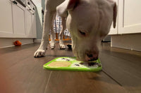 An image of a white dog standing over a green lick mat. The dog is licking the mat which has yogurt and berries.