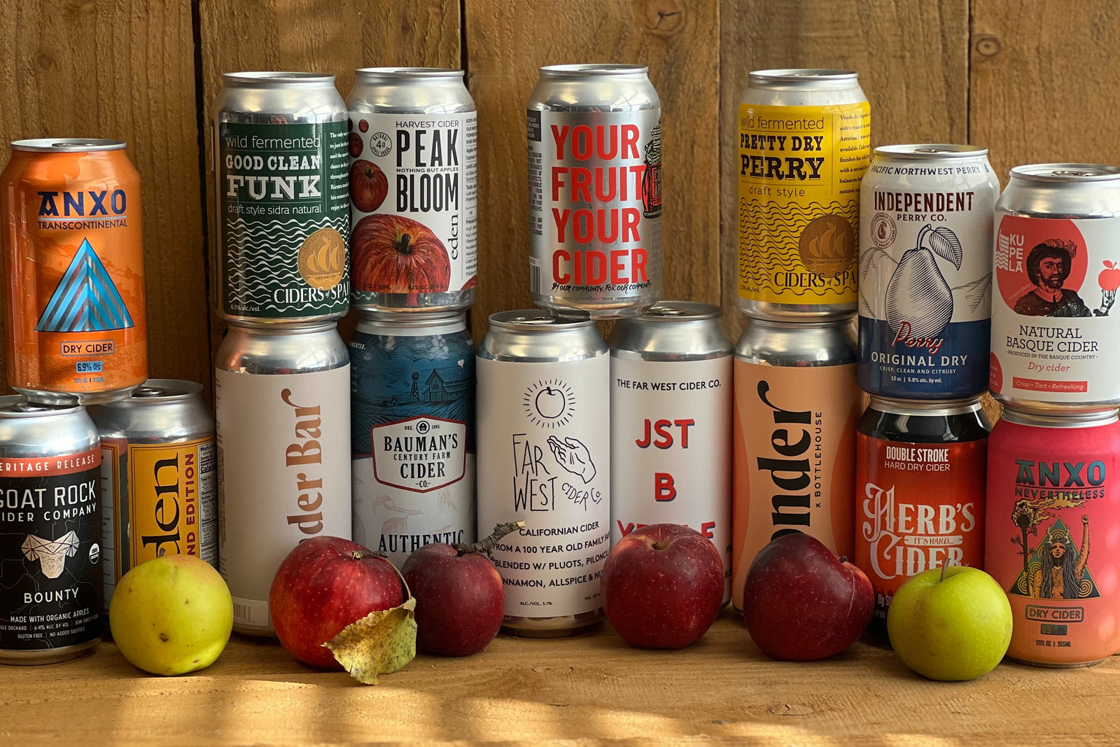 A mix of 12 various cans of cider surrounded by apples in front of a wood fence. 