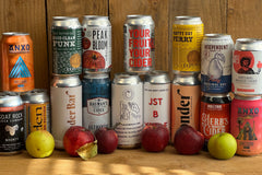 A mix of 12 various cans of cider surrounded by apples in front of a wood fence. 