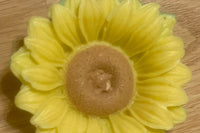 A sunflower-shaped and -colored soap rests on a woodgrain-patterned floor; it is approximately four inches across.