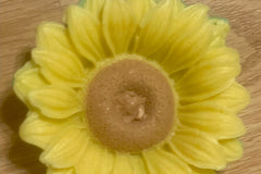A sunflower-shaped and -colored soap rests on a woodgrain-patterned floor; it is approximately four inches across.