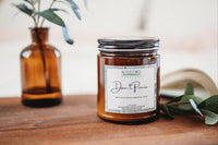 a candle named don't panic sits on a brown tabletop alongside an open book and some plants and plant cuttings.