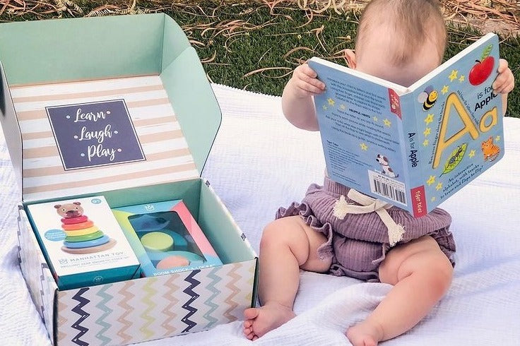 Baby reading an alphabet book while sitting on blanket next to a baby discovery box month 12 containing toys and puzzles for a 1 year old. 