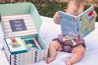 Baby reading an alphabet book while sitting on blanket next to a baby discovery box month 12 containing toys and puzzles for a 1 year old. 