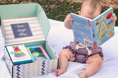 Baby reading an alphabet book while sitting on blanket next to a baby discovery box month 12 containing toys and puzzles for a 1 year old. 