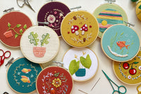 Overhead photo of 10 embroidery projects of varying colors, designs, and textures, piled on top of one another amongst scattered supplies.