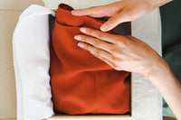 Two hands are carefully removing a dark orange piece of clothing from a box. Box has tissue paper in it. 
