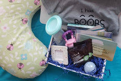 A shirt and nursing pillow next to a subscription box filled with bath bombs, protein mix, affirmation cards, and a frame.