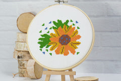 orange and yellow flower with green leaves on white embroidery fabric inside wood hoop beginner friendly counted cross stitch craft