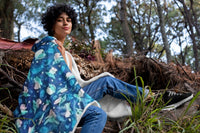 Lady in a forest with a Blue, Green, and White fuzzy Baby Yoda blanket wrapped around her shoulders and back.         