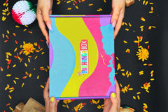 Pachanga Box featuring two hands grabbing on the the Pachanga Box with a black background with flower pedals. 