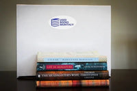 A stack of used books in front of a Used Books Monthly subscription box showcasing a book subscription service